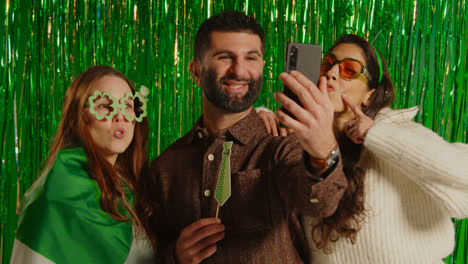 Studio-Shot-Of-Friends-Dressing-Up-With-Irish-Novelties-And-Props-Posing-For-Selfie-Celebrating-St-Patrick's-Day-Against-Green-Tinsel-Background-2